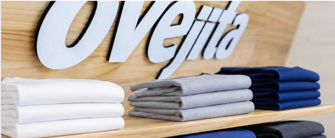 Letras blancas de Ovejita sobre madera, con franelas dobladas blancas, grises y azules.